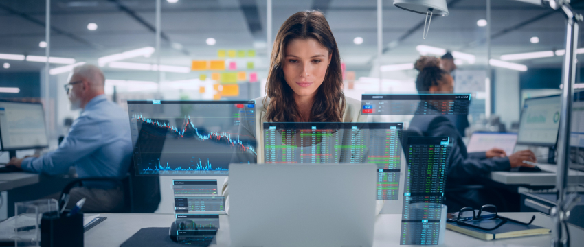 Uma mulher está sentada a uma mesa em um escritório moderno, concentrada em seu laptop, enquanto gráficos de negociação Forex e gráficos de dados são projetados à sua frente. Outras pessoas trabalham em mesas ao fundo.