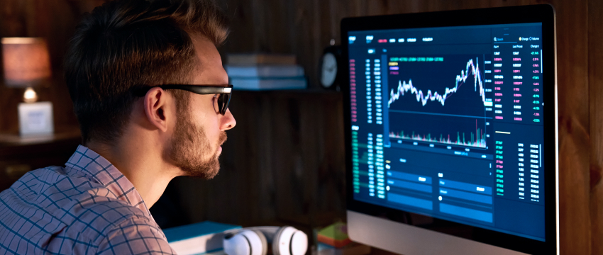 A man in glasses focuses intently on a computer screen displaying financial stock charts in a dimly lit room, conveying concentration and analysis.
