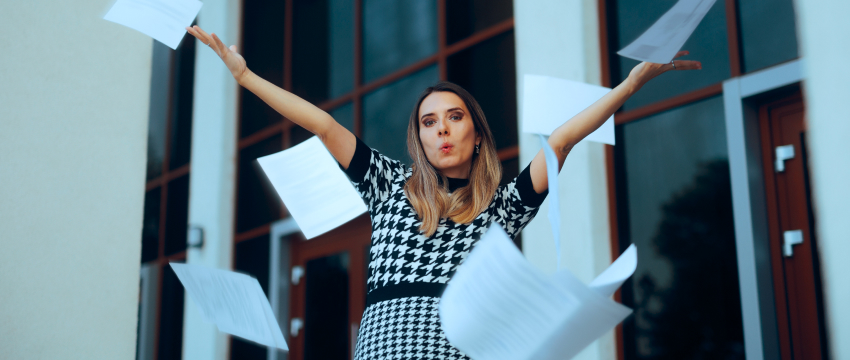 A forex trader woman in a black-and-white patterned dress joyfully throws papers into the air outside a modern building in trading plan