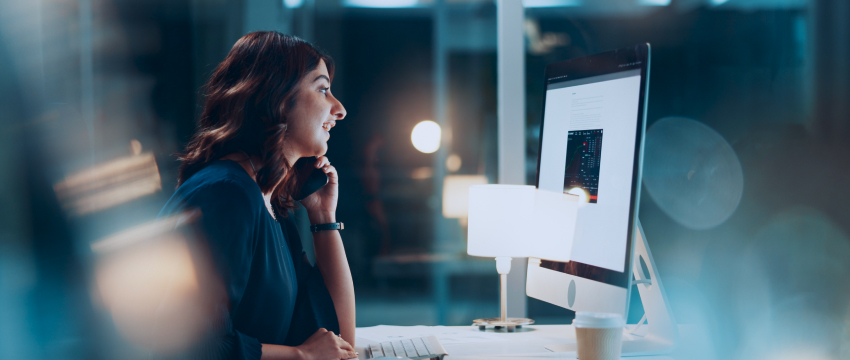 A woman is seated at a desk, engaged with her computer screen, managing her forex trading and daily life balance.