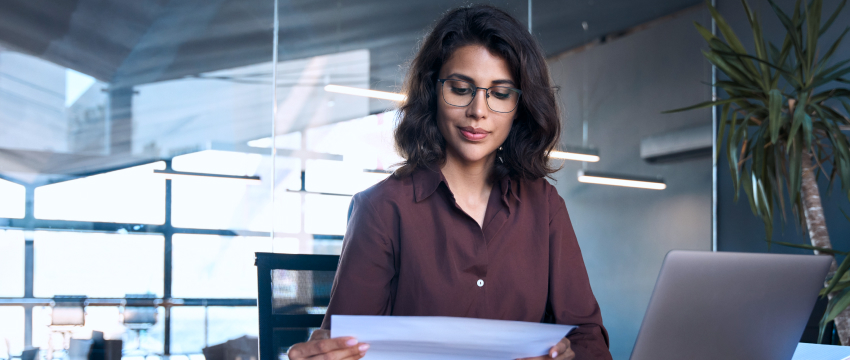 A woman with glasses reviews a piece of paper, strategizing her forex trading plan while working on her laptop.